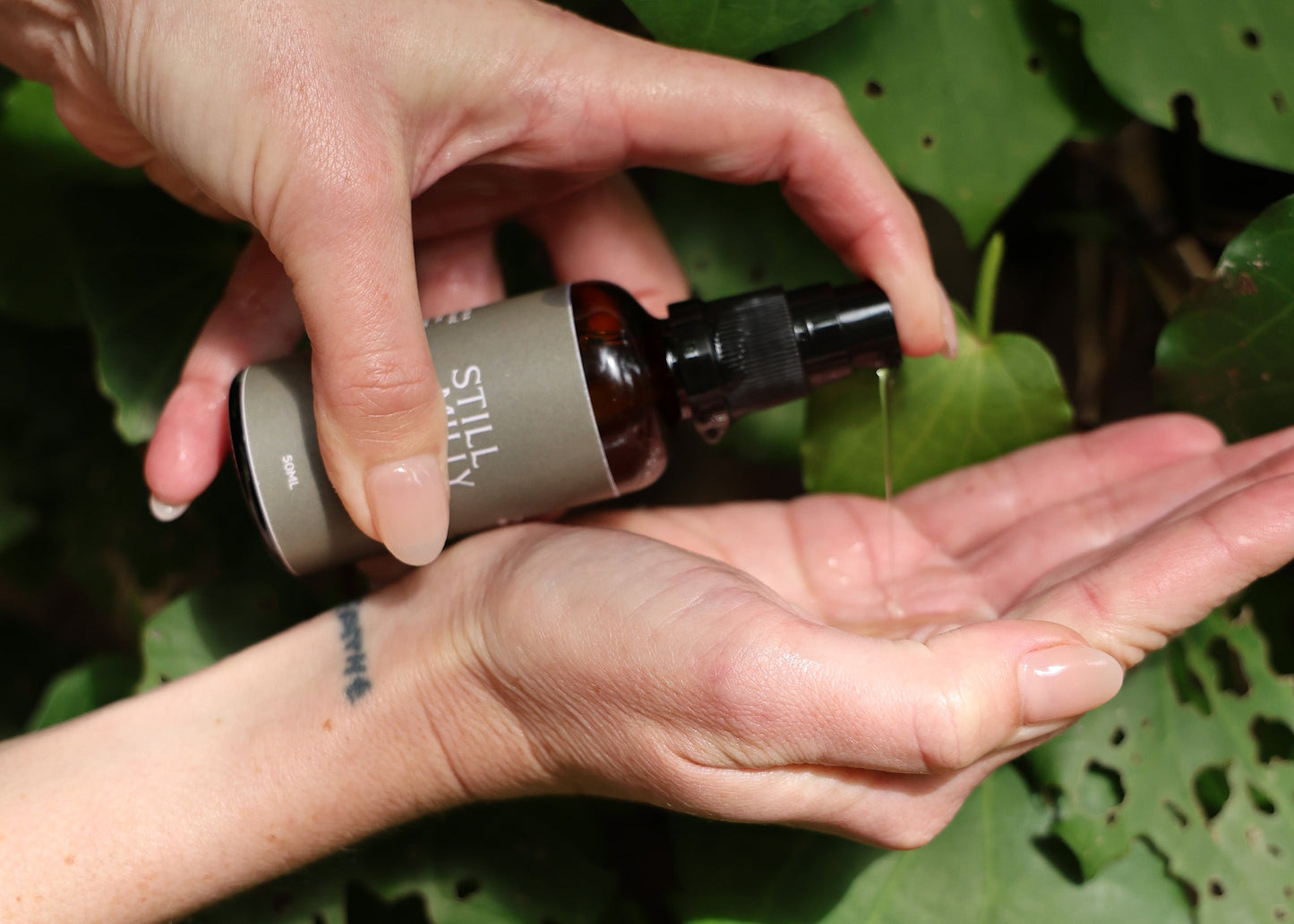 Hands dispensing Still Milly Kānuka and Mandarin Cleanser with Kawakawa in background