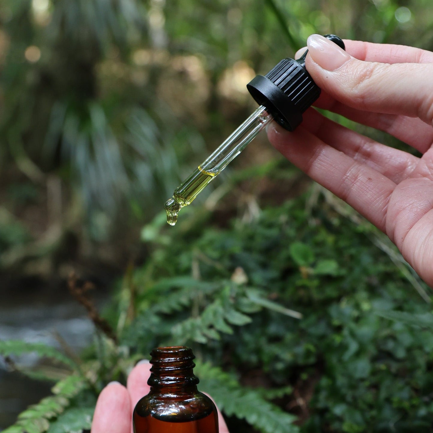 Hand holding a dropper with a liquid over a small bottle against a natural background